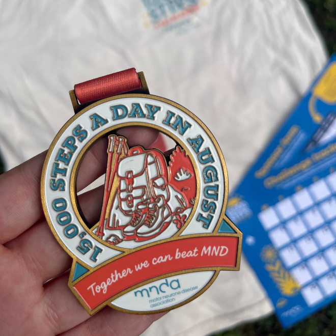 a hand holding the steps medal, it says 15,000 steps a day in august in clue writing and there is an image of a backpack in the middle