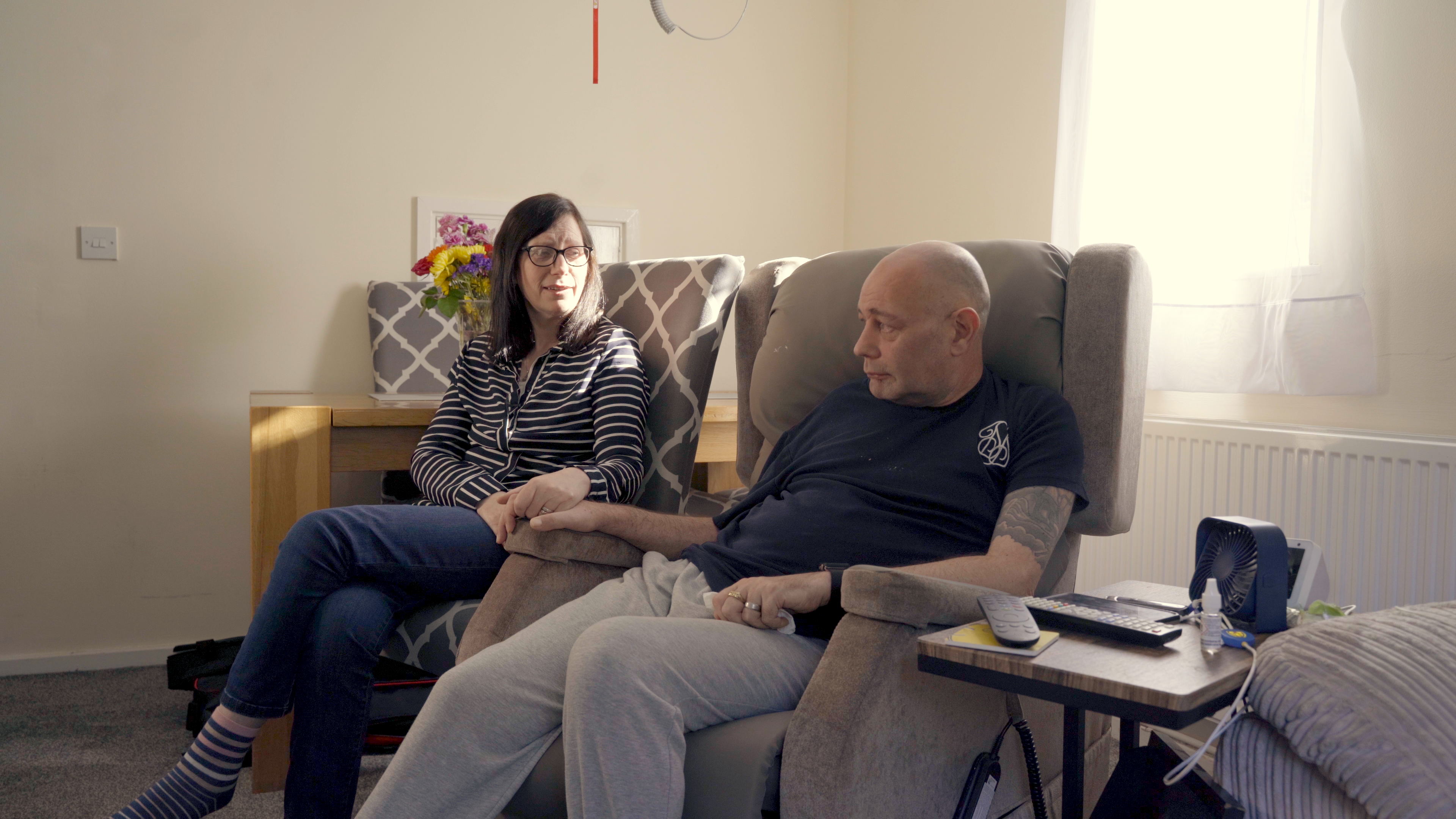 A man with MND sat on a riser recliner chair holding hands with a woman sat next to him